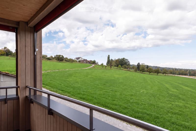 Balcony with wooden railings overlooking a large, green lawn and distant houses under a partly cloudy sky.