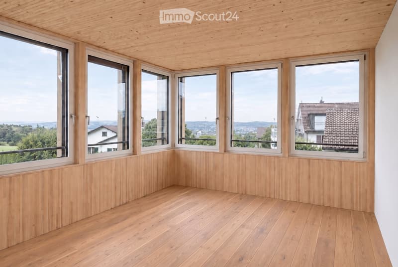 Empty room with wooden flooring and walls, large windows offering a scenic view of the landscape and neighboring houses.