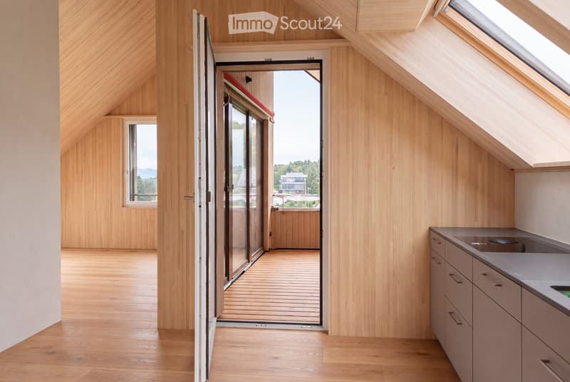 Bright, wooden-floored room with a kitchen area, large windows, and a sliding glass door leading to a wooden deck with a scenic view.