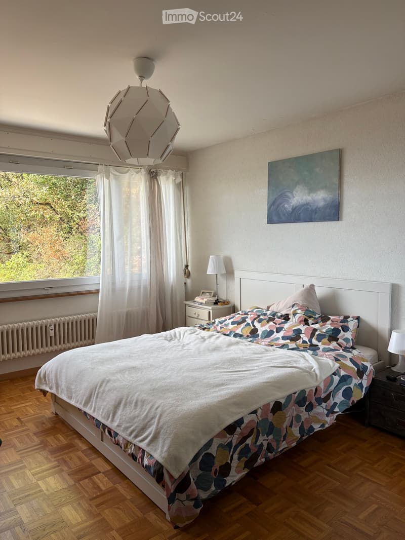 A well-lit bedroom with a large bed, colorful bedding, a nightstand with a lamp, a painting on the wall, and a window with curtains.
