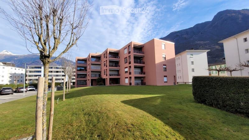 Modern multi-story residential building with pink facade and balconies, surrounded by green lawn and trees, with mountains in the background.
