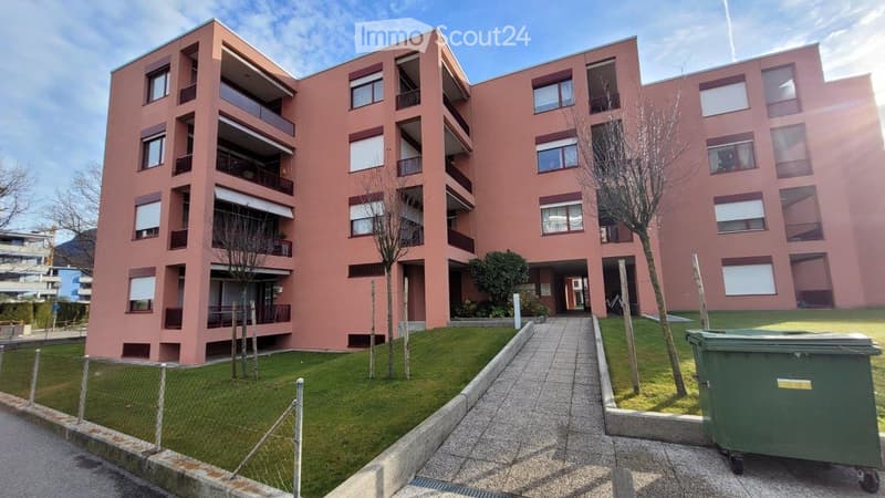 Modern multi-story apartment building with pink facade, multiple balconies, and surrounding greenery.