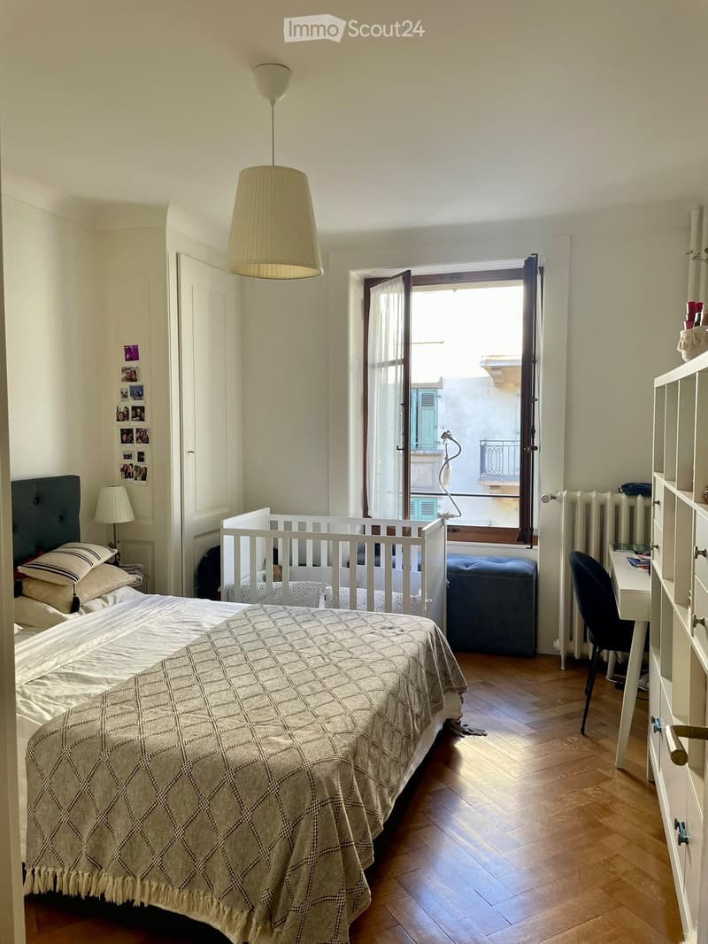 Cozy bedroom with a large bed, nightstand, baby crib, desk, and balcony access. Features wooden flooring and white walls.