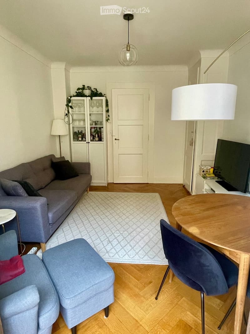 A modern living room with a grey sofa, a blue armchair, a round wooden table with chairs, a white cabinet, and a TV screen. The room features wooden flooring and a patterned rug.