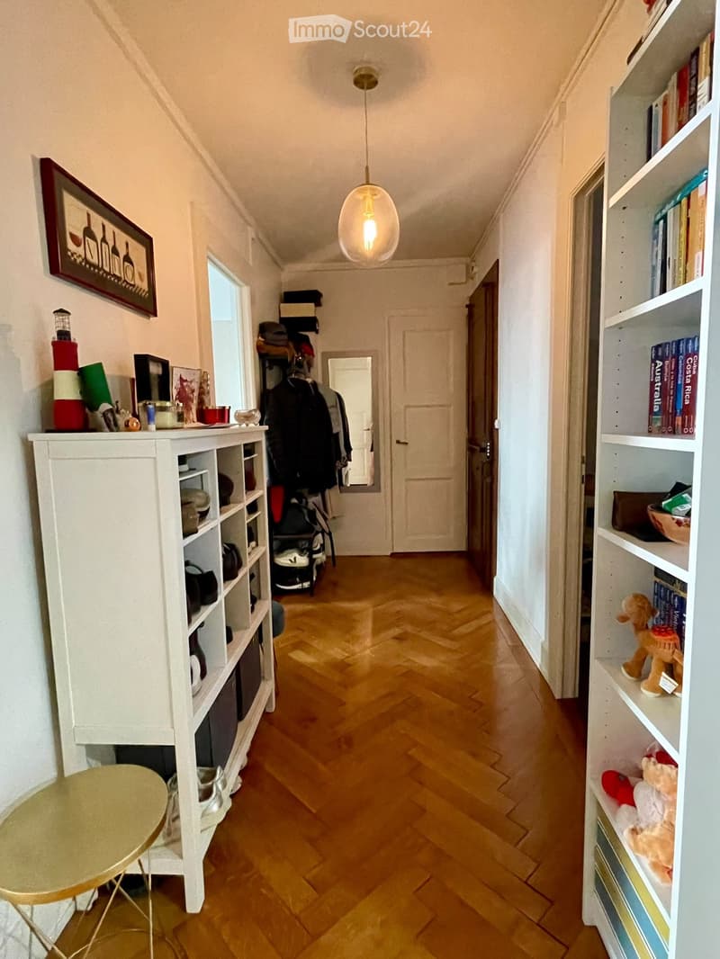 A narrow hallway with wooden flooring, white walls, and built-in shelving units on both sides. The left side features a small round table, a coat rack with jackets, and a framed picture. The right side has bookshelves with books, toys, and decorative item