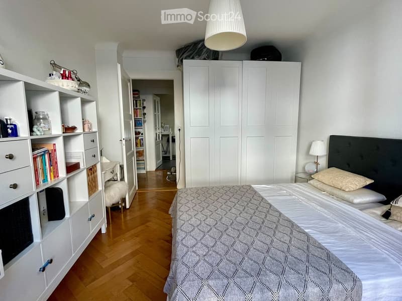 A well-lit bedroom with a wooden floor, a large bed with a grey headboard and patterned blanket, a bedside table with a lamp, a large white wardrobe, and a built-in shelf unit with books and decorative items.