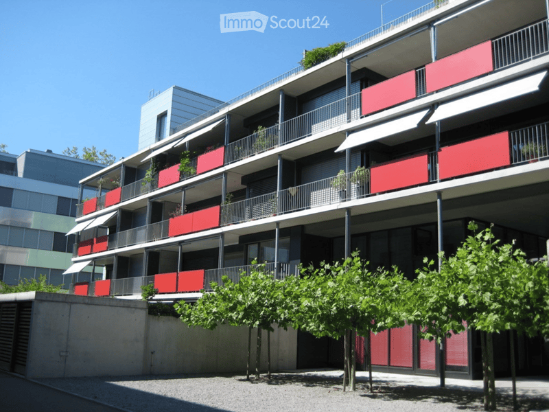 Modern multi-story residential building with red balconies, metal railings, and greenery on the ground level.