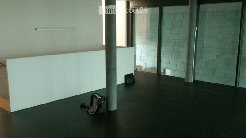 Empty room with dark flooring, white walls, and large glass windows. Minimal furnishings, including a small black bag and a chair.