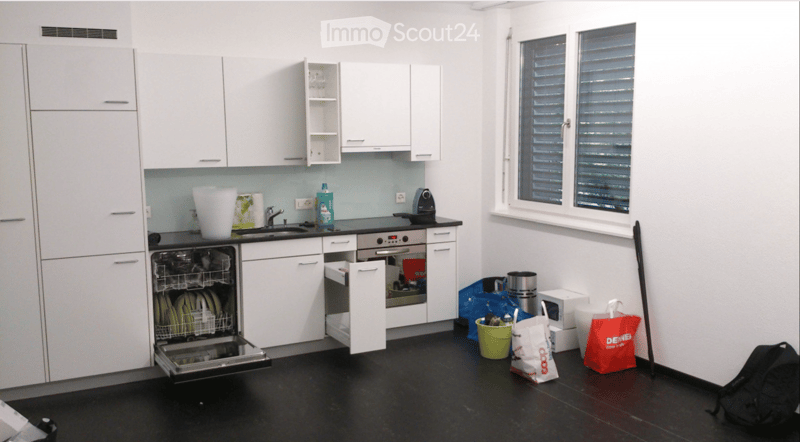 Modern kitchen with white cabinets, black countertops, and stainless steel appliances. Features a dishwasher, oven, sink, and various items including a backpack and handbags.
