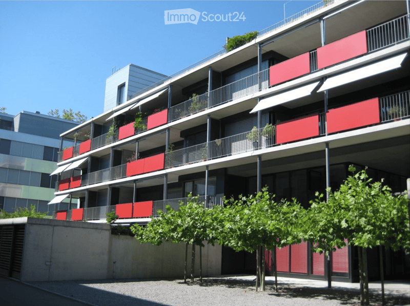 Modern multi-story residential building with red balconies, metal railings, and greenery in front.