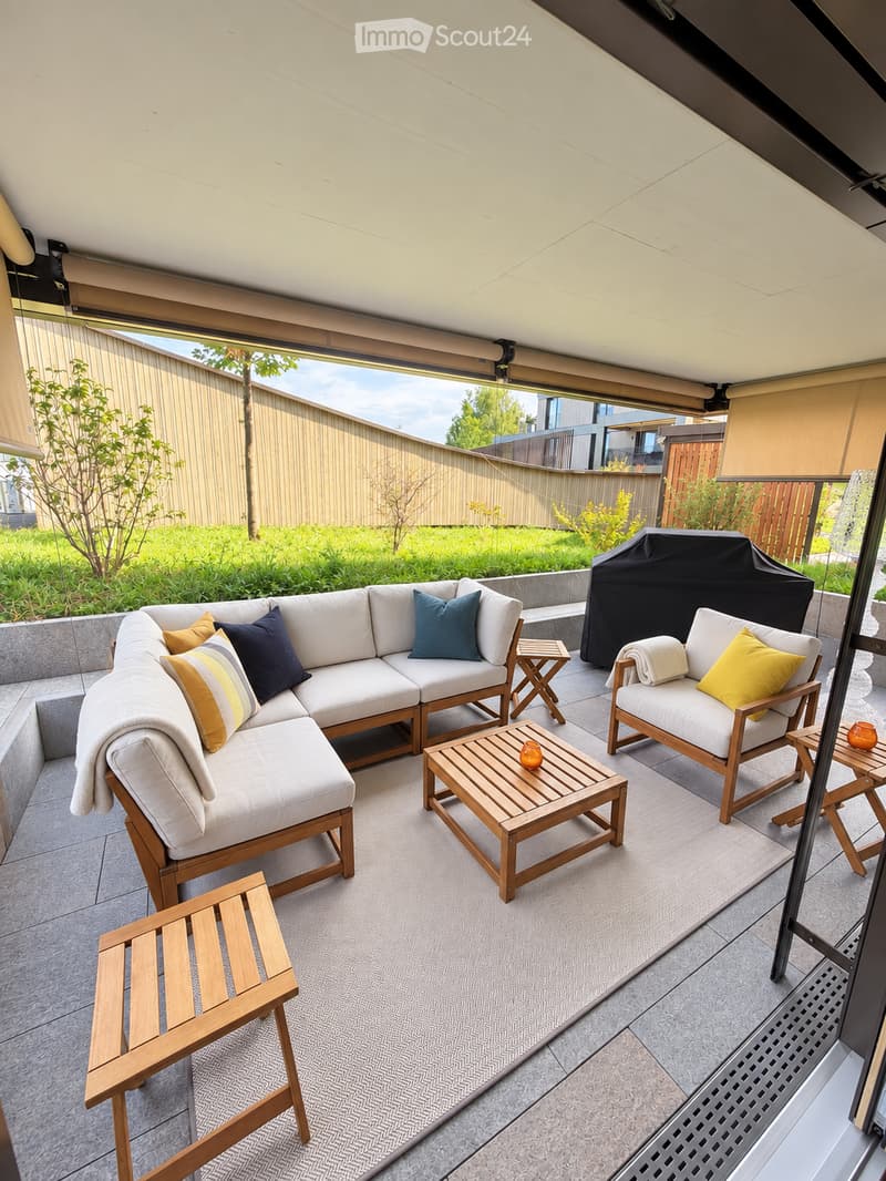 A modern outdoor patio with a white sectional sofa, wooden coffee table, and matching chairs, all set on a grey tiled floor with a grey rug.
