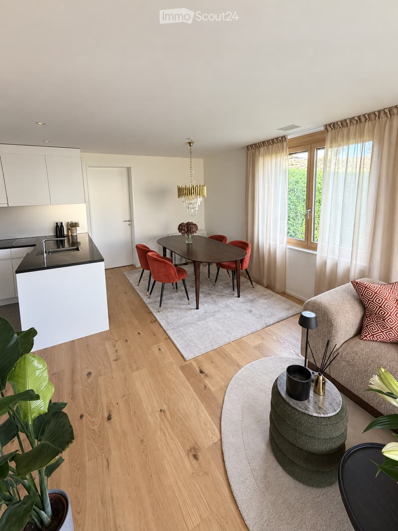 Open-plan living and dining area with light wooden flooring, a modern sofa, a coffee table, a dining table with chairs, a kitchen counter, and a chandelier.
