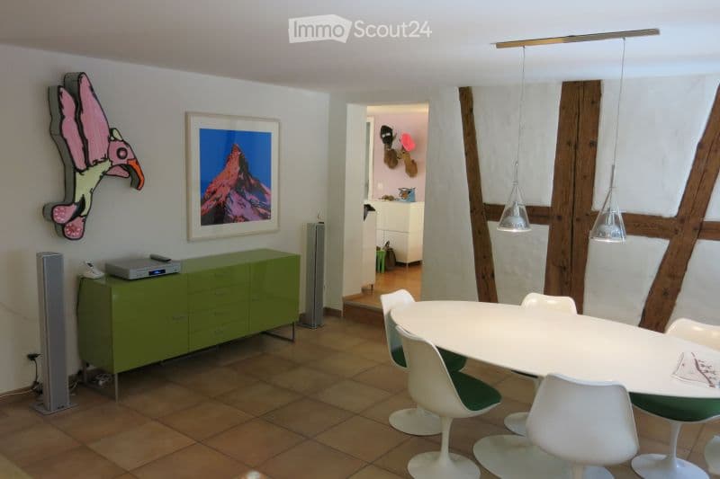 Modern living and dining area with a green cabinet, white dining table with chairs, pendant lamps, and wall art including a framed mountain scene and a colorful bird sculpture.