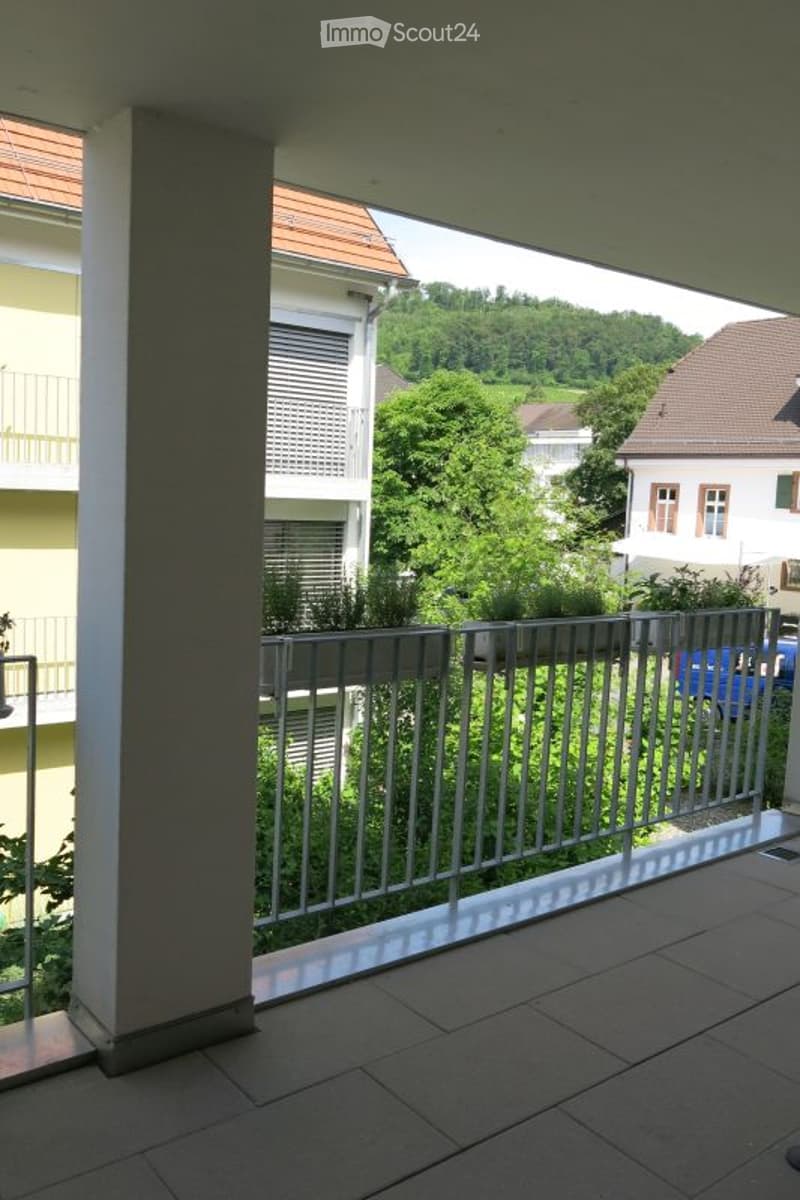 Balcony with tiled floor, metal railing, and potted plants, overlooking a green area with trees and neighboring buildings.