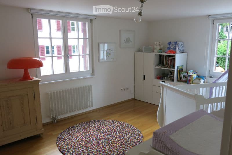 Bright, child-friendly bedroom with wooden flooring, large windows, a white bed with purple bedding, a colorful round rug, a white cabinet, a bookshelf filled with books and toys, and an orange lamp on a wooden dresser.