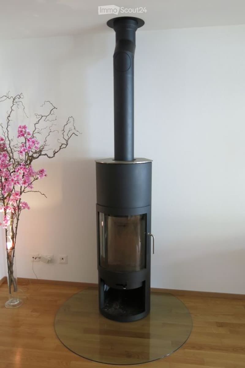 Modern black wood-burning stove with a tall chimney, placed on a glass platform in a room with wooden flooring and white walls. A vase with pink flowers is visible to the left.