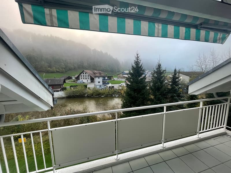 Balcony with white railing and tiled floor, overlooking a river and misty mountains with houses and trees.