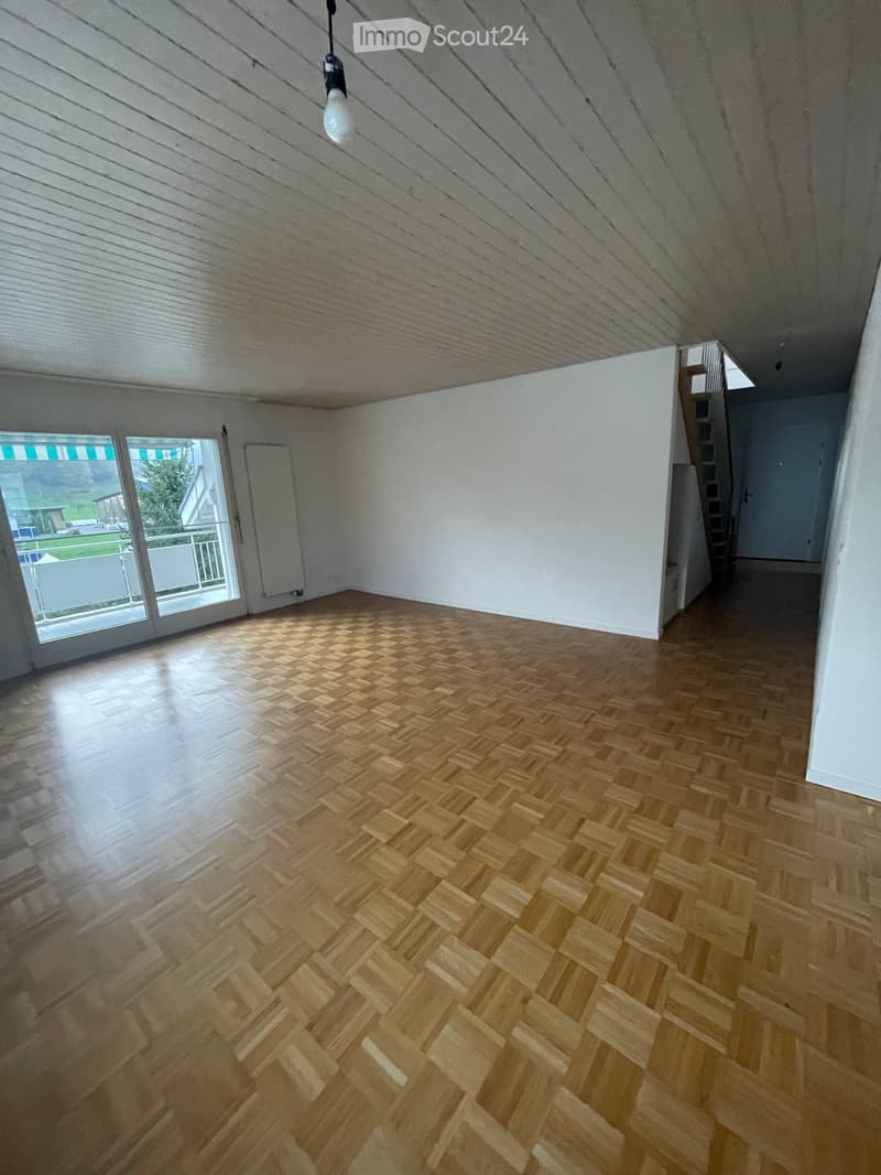 Empty room with wooden herringbone flooring, white walls, and a large window with a view of the balcony and outdoor area.
