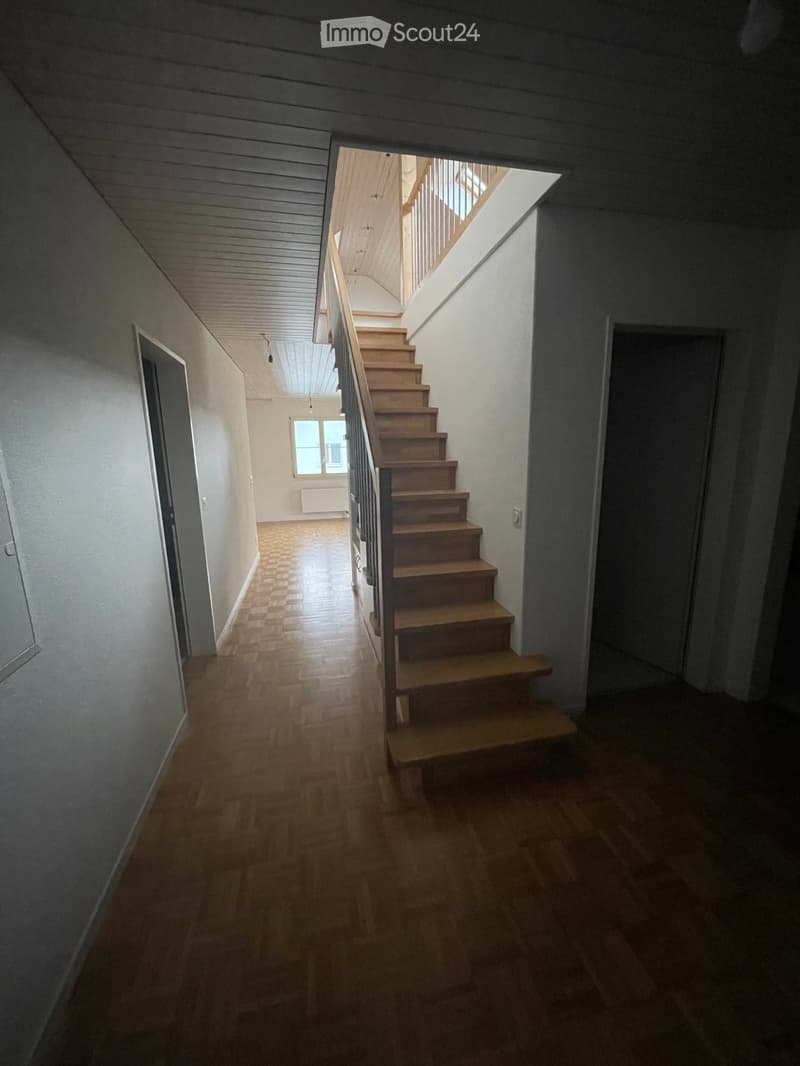Empty hallway with wooden flooring, white walls, and a staircase leading upwards with wooden steps and a metal railing.