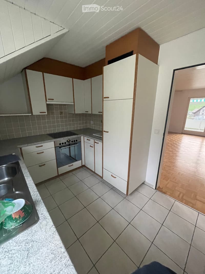 Modern kitchen with white cabinets, a refrigerator, an oven, and a granite countertop. The floor is tiled, and there is a doorway leading to another room with wooden flooring.
