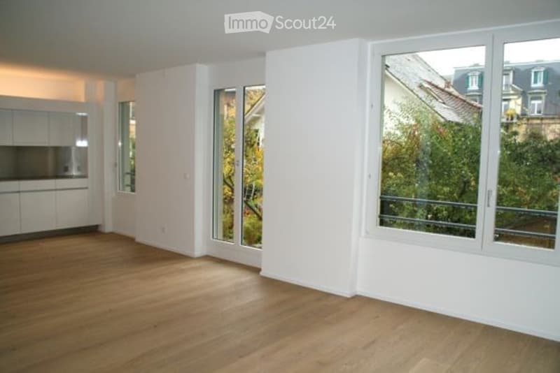 Empty room with wooden flooring, white walls, large windows, and a small kitchen area visible in the background.