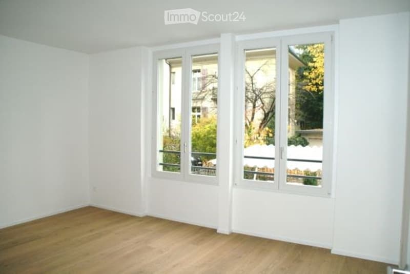Empty room with wooden flooring and large windows, showing a view of the outside area with trees and a building.
