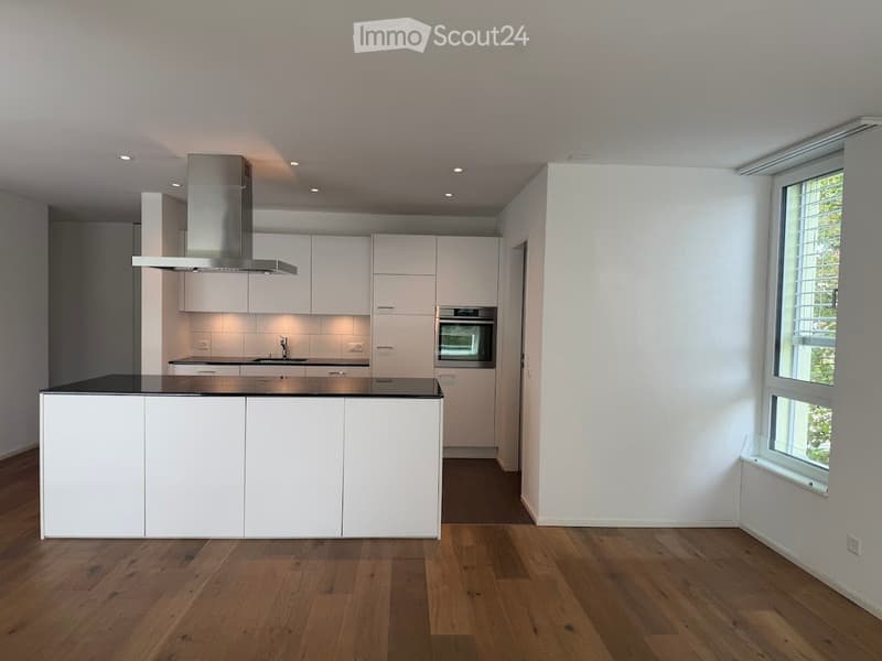 Modern, minimalist kitchen with white cabinets, a central island, and integrated appliances including an oven and range hood. The space features wooden flooring and large windows allowing natural light.