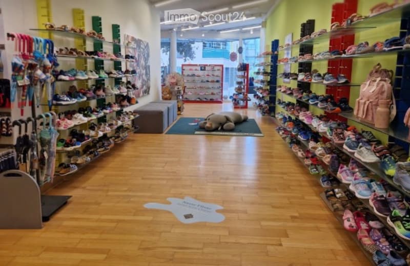 A brightly lit shoe store with wooden flooring, shelves filled with various shoes, a plush toy on the floor, and a backpack on display.