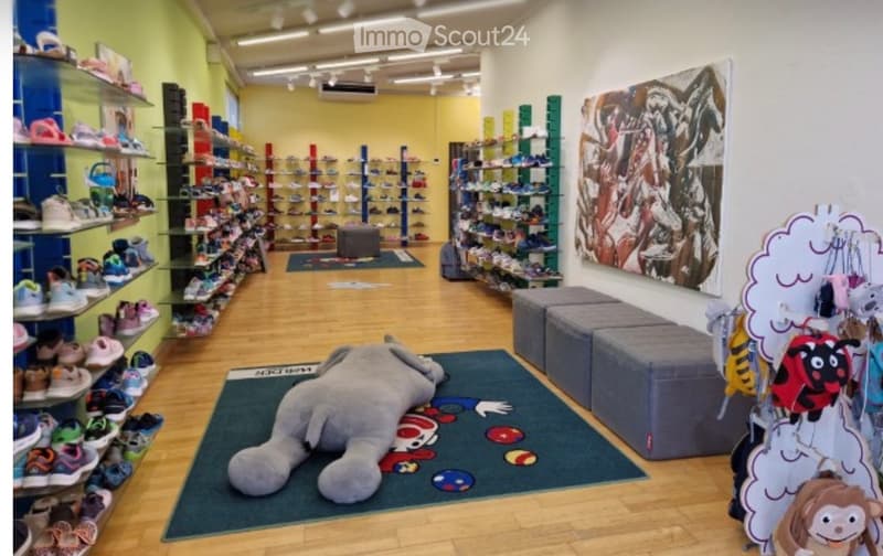 A brightly lit children's shoe store with wooden flooring, shelves filled with shoes, a large plush toy, and colorful decor including a cloud-shaped display and a large picture on the wall.