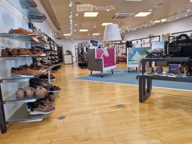A well-lit shoe store with wooden flooring, glass shelves displaying various shoes, a small seating area with a sofa, table, and lamp, and additional display items including handbags and a mirror.