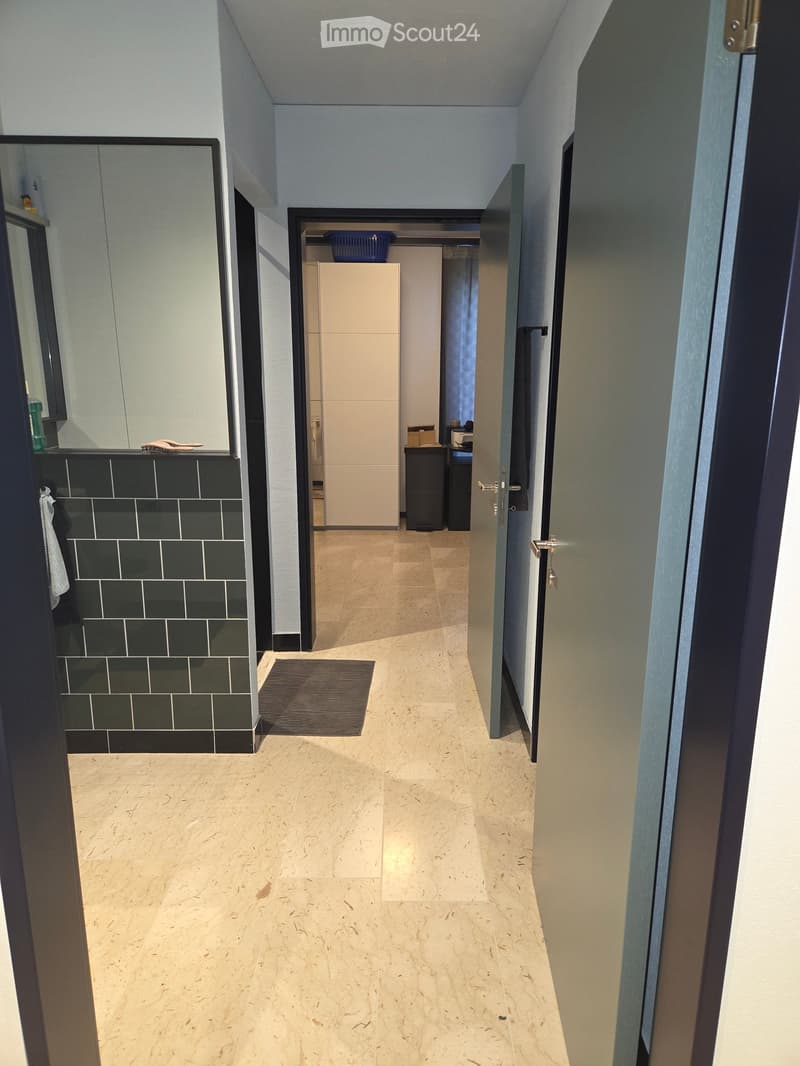 Clean, modern hallway with light-colored tiled flooring, white walls, and several doors leading to different rooms. A small rug is placed in the center of the hallway.