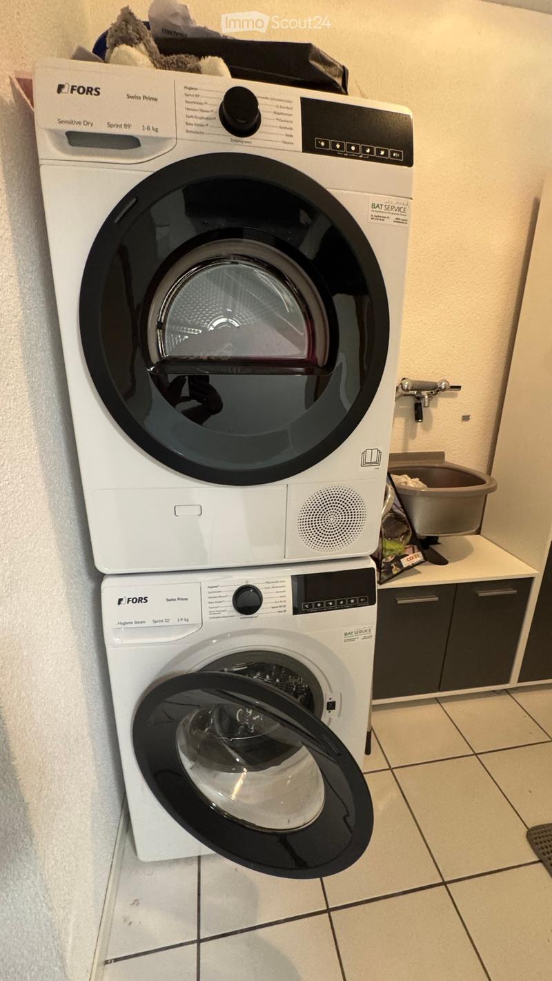 A modern bathroom with a stacked washing machine and dryer unit, control panels, and a small shelf with a bucket and other items.