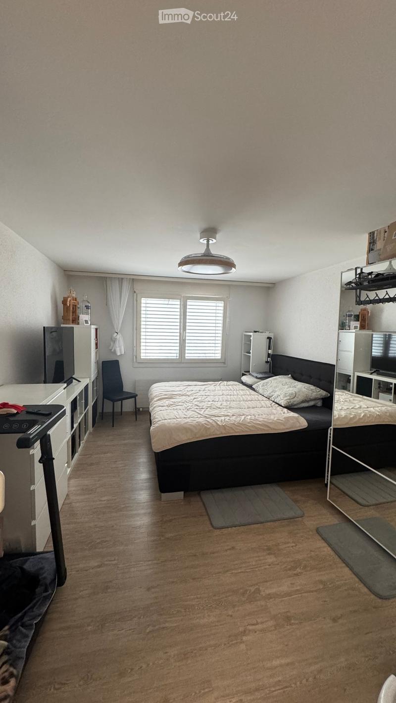 A modern bedroom with a large bed, a chair, a mirror, shelves, and a lamp. The room features wooden flooring and large windows with curtains.