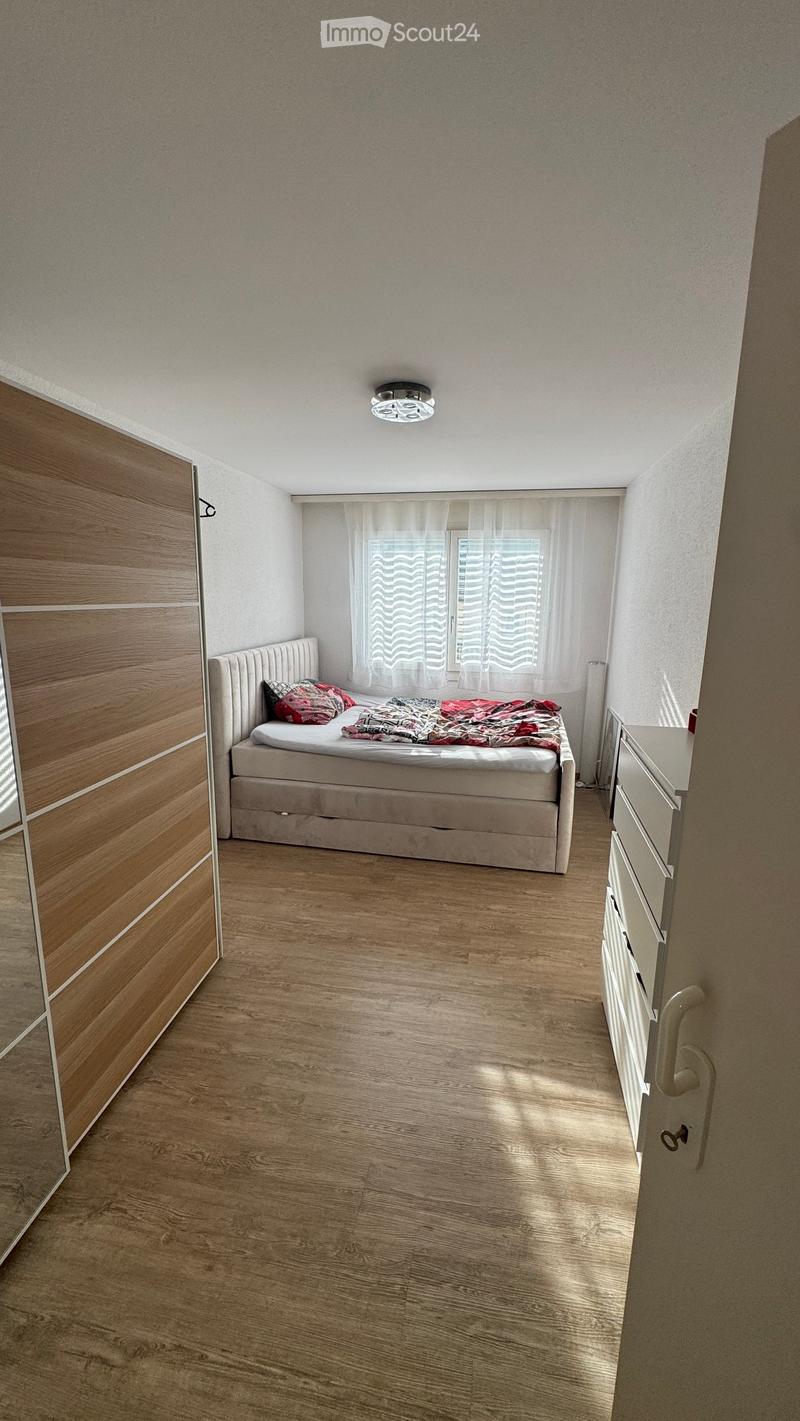 Cozy bedroom with wooden flooring, a bed with a headboard, a nightstand, and a wardrobe. The room features a window with white curtains and a ceiling light fixture.