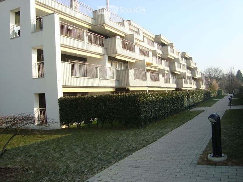Modern multi-story apartment building with multiple balconies and a neatly trimmed hedge line along a paved walkway.