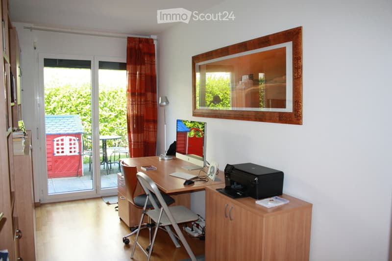 Bright, modern home office with wooden flooring, large window with view, desk with computer, chair, printer, and cabinet. Mirror on wall, red curtain, and outdoor seating visible through sliding glass door.