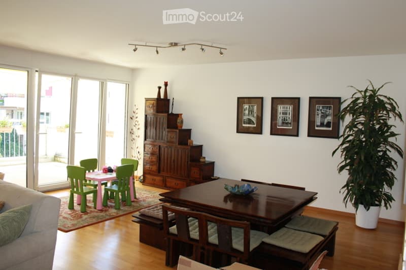Bright living and dining area with large windows and sliding doors leading to a balcony. Features a grey sofa, coffee table, dining table with chairs, wooden cabinet, decorative vases, picture frames, and a large potted plant.
