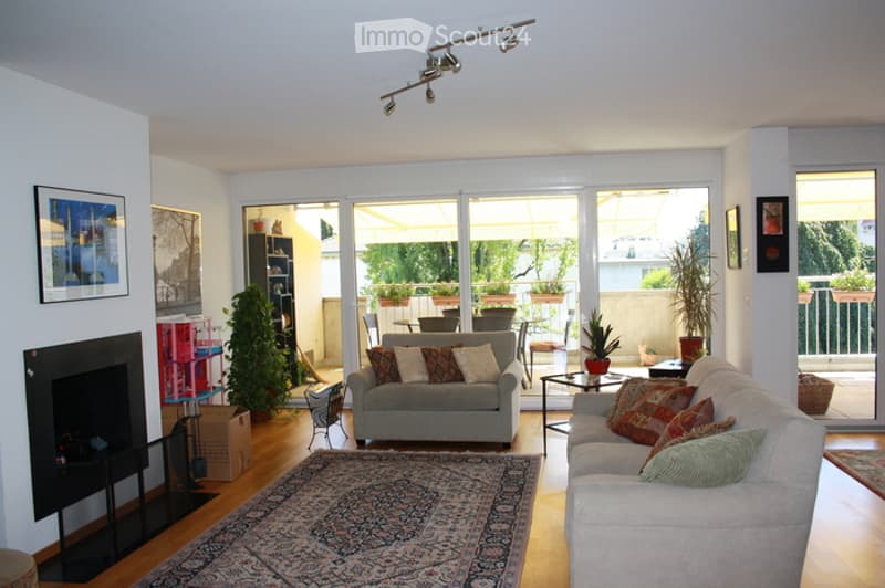 Bright living room with large sliding glass doors leading to a balcony, fireplace, two sofas, coffee table, bookshelf, and decorative plants.