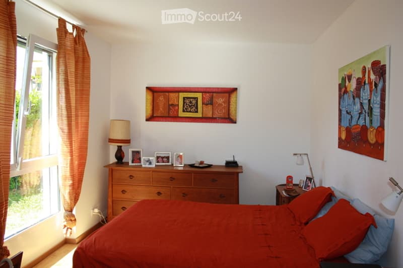 Bright bedroom with a large bed, wooden dresser, nightstand, and artwork on the walls.