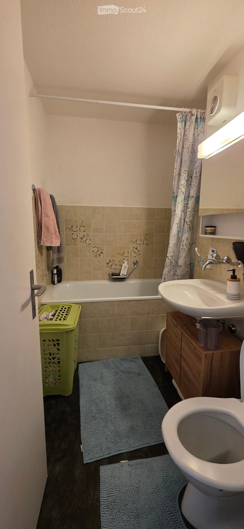 Bathroom with a bathtub, sink, toilet, and wooden cabinet. Features a tiled wall, shower curtain, and two blue rugs on the floor.