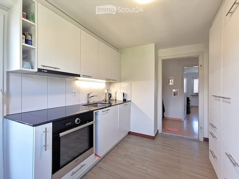Modern kitchen with white cabinets, stainless steel appliances, and a wooden floor. The kitchen features an oven, sink, and faucet, with a doorway leading to another room.