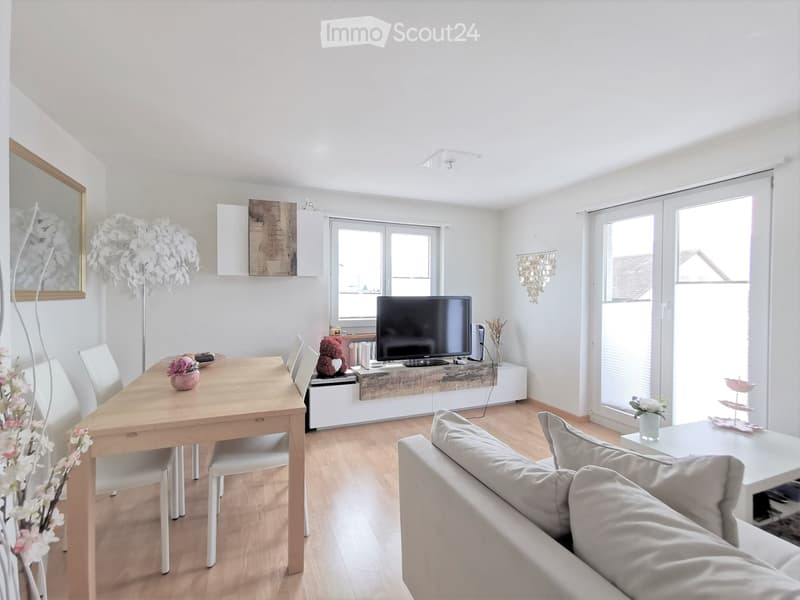 Bright, modern living and dining area with white walls, light wood flooring, and large windows. Features a white sofa, coffee table, dining table with chairs, TV on a wooden stand, and decorative items including a floor lamp and vases.