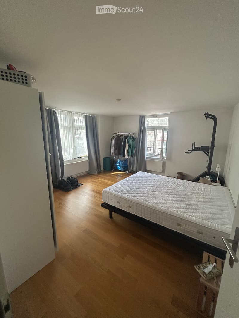 A furnished bedroom with a wooden floor, a bed with a mattress, a clothing rack, a small stool, and a lamp near the window.