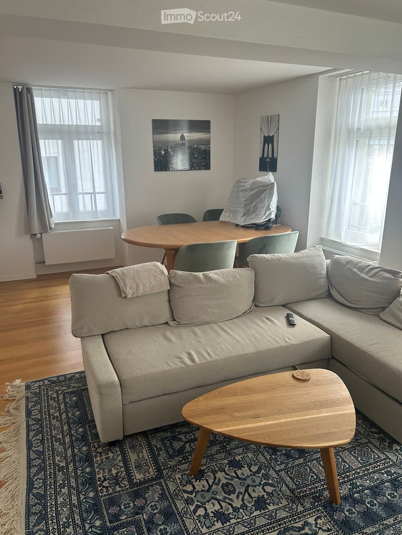 A modern living room with a grey sectional sofa, wooden coffee table, dining table with chairs, and large windows with sheer curtains.