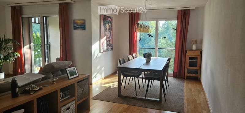 Bright living and dining area with wooden flooring, large windows offering a scenic view, a sofa, dining table with chairs, a side cabinet, and decorative items.
