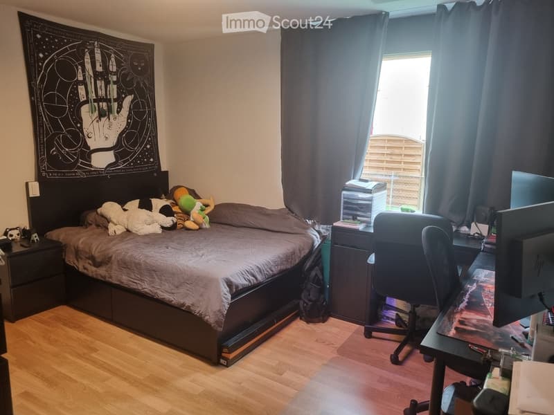 A cozy bedroom with a large bed, a desk with a computer, a chair, and various decorative items including a poster and toys.