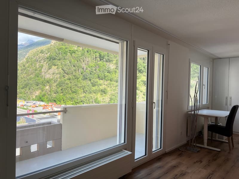 Bright room with large windows and a balcony overlooking a green, forested mountain landscape. Includes a white table, black chair, and a modern floor lamp.