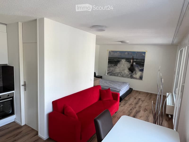 A compact, furnished room with a red sofa, a single bed, a small table and chair, an oven, and a large picture frame on the wall.