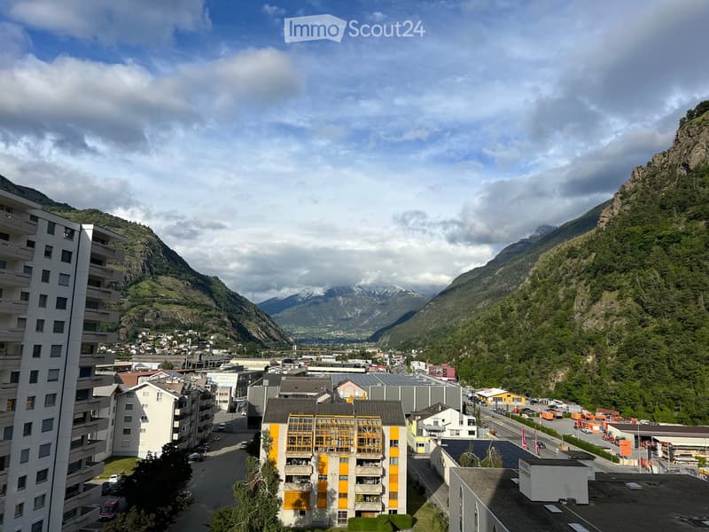 A panoramic view of a town nestled in a mountainous valley, with modern apartment buildings, roads, and surrounding mountains.