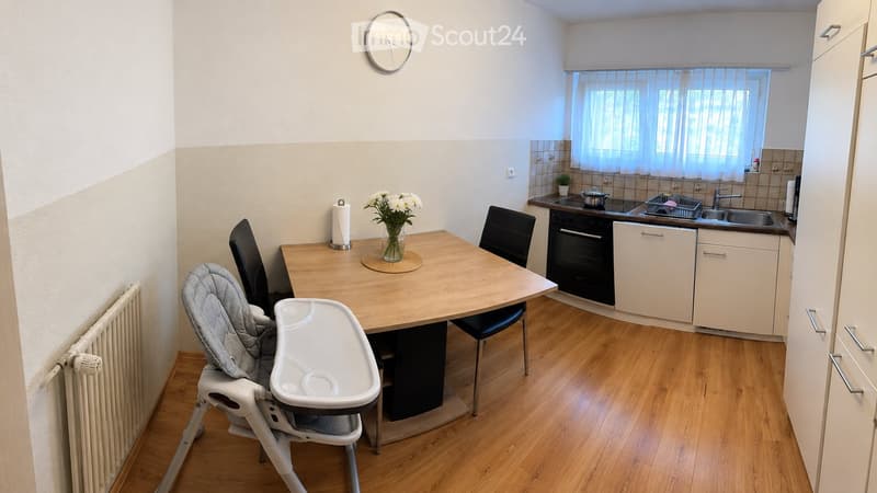 Modern kitchen with light wooden flooring, white cabinets, a dining table with chairs, a baby chair, and a vase of flowers. The kitchen features an oven, sink, and a window with a curtain.